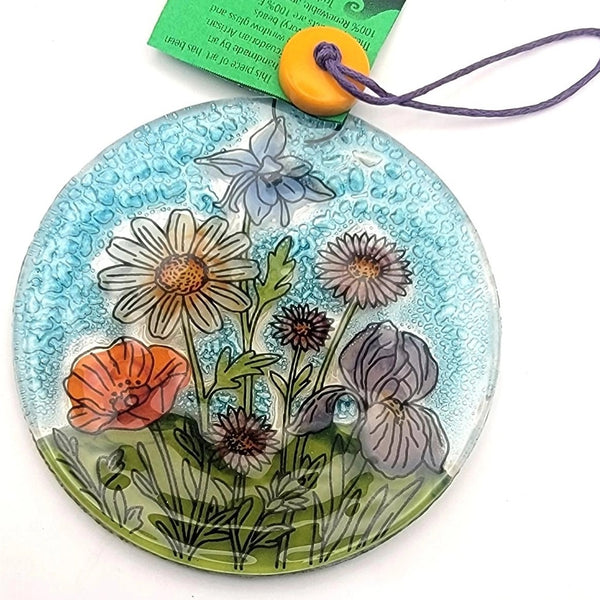 A round glass pendant ornament featuring a hand-painted floral design with daisies, a poppy, and purple flowers on a textured light blue background. The pendant has a purple hanging cord and an orange bead, with a green product tag attached.