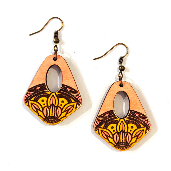 A pair of wooden dangle earrings with a natural wood top half, a central oval cutout, and a lower half featuring a yellow, orange, and black mandala pattern, hanging from bronze-colored fishhook wires on a white background.