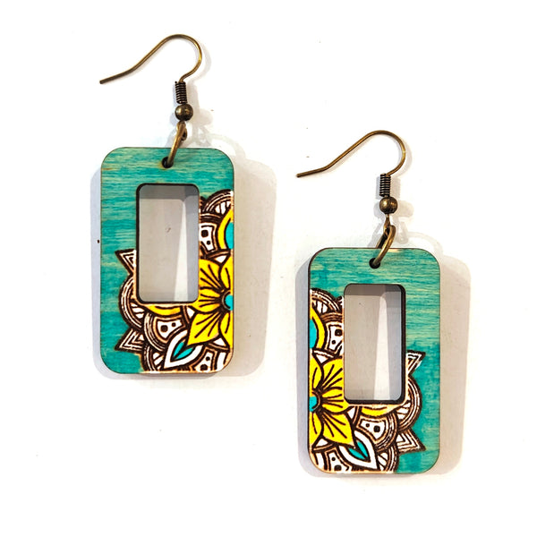 A pair of rectangular drop earrings with a turquoise wood-grain finish, featuring a central cutout and a yellow mandala floral pattern on the lower half, hanging from bronze-colored fishhook wires on a white background.