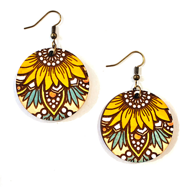 A pair of round drop earrings with a hand-painted mandala sunflower design in yellow, brown, white, and teal, hanging from antique bronze fishhook wires on a white background.