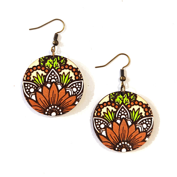 A pair of round dangle earrings with a colorful floral mandala pattern on a cream background, featuring rust orange, lime green, and black details, suspended from bronze-colored fishhook wires on a white background.