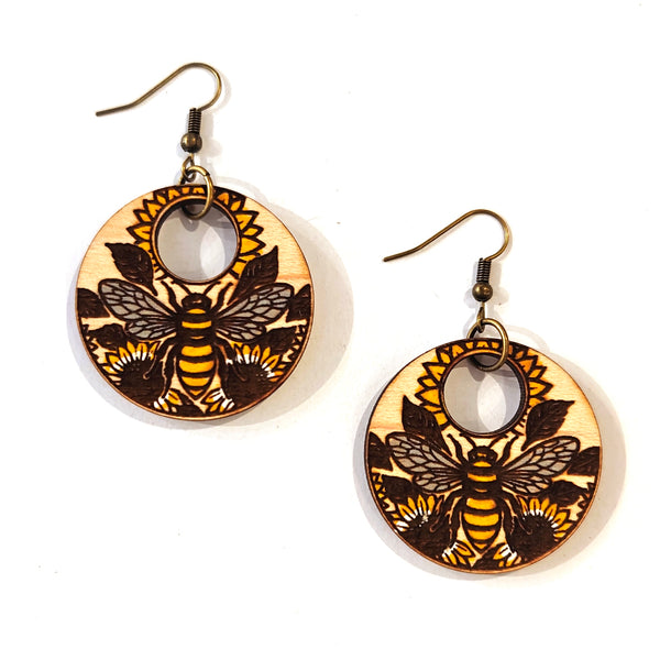 A pair of round wooden dangle earrings featuring a detailed laser-burned bee and sunflower design in yellow, black, and natural wood tones, suspended from antique bronze fishhook wires on a white background.