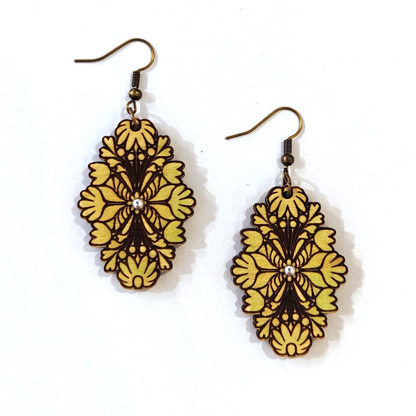 A pair of dangling earrings with ornate, symmetrical floral cutout designs in yellow and dark brown, suspended from bronze-toned fishhook wires on a white background.