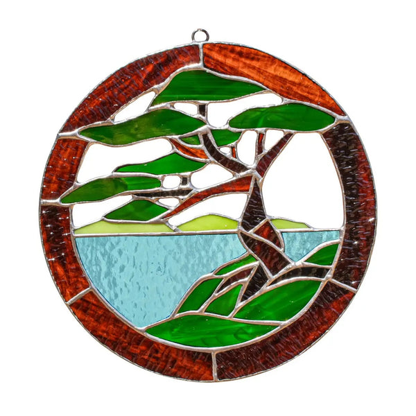 A circular stained glass window panel featuring a stylized green tree with a brown trunk, set against a light blue water background and a reddish-brown border.