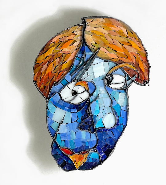 A mosaic portrait wall art piece depicting a face with blue-toned tiles, orange and brown hair, white eyes, and small red and yellow accents.