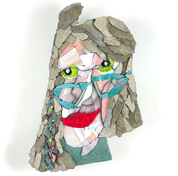 A mosaic portrait wall art piece featuring a stylized face made from cut glass, stone, and paper fragments. The artwork includes bright green eyes, turquoise glasses, red lips, and a gray stone-textured hair area.