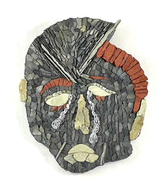 A  mosaic wall art piece depicting a crying face, constructed from gray slate tiles, terracotta orange stones, cream-colored stones, and small metallic silver chains.
