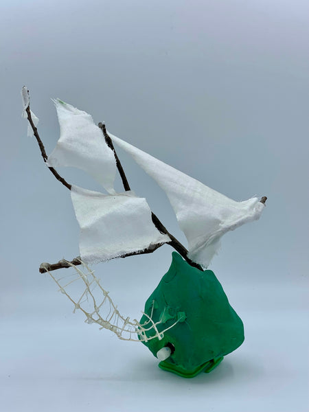 A handmade decorative sailing ship sculpture made from a green clay-like base, brown branches, and white fabric sails, photographed against a plain light gray studio background.
