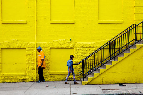 Photograph of two people standing against a bright yellow wall with a black metal staircase.