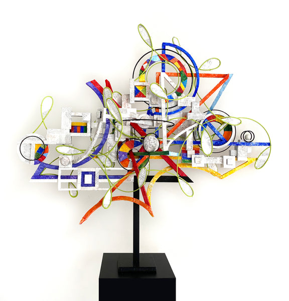 Abstract metal sculpture on a black pedestal stand, featuring layered geometric shapes, circles, lines, and colored panels in blue, red, yellow, green, orange, and white against a plain white background.