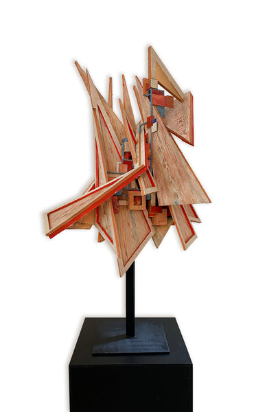Abstract wooden sculpture with layered triangular and angular wood panels in natural wood tones and red painted edges, mounted on a black metal pole and displayed on a black pedestal against a white background.