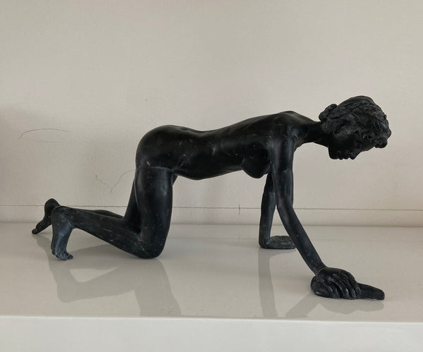 A black clay sculpture of a nude female figure in a crawling position mopping the floor, displayed on a white shelf against a light beige wall.