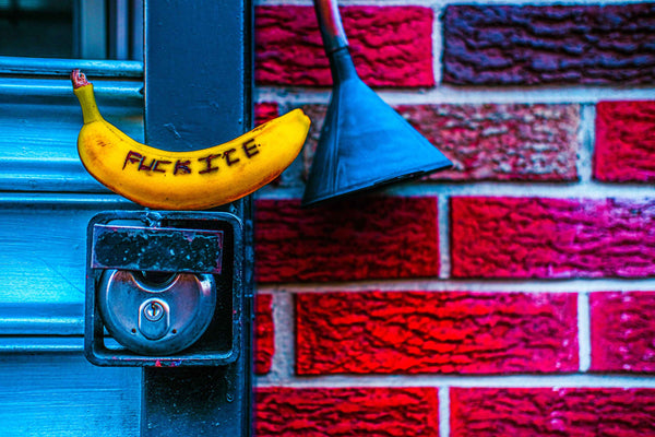 Photograph of a banana with text attached to a door handle against a red brick wall