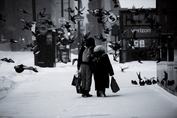 Two people walking on a snowy street with birds flying around, black and white photo.